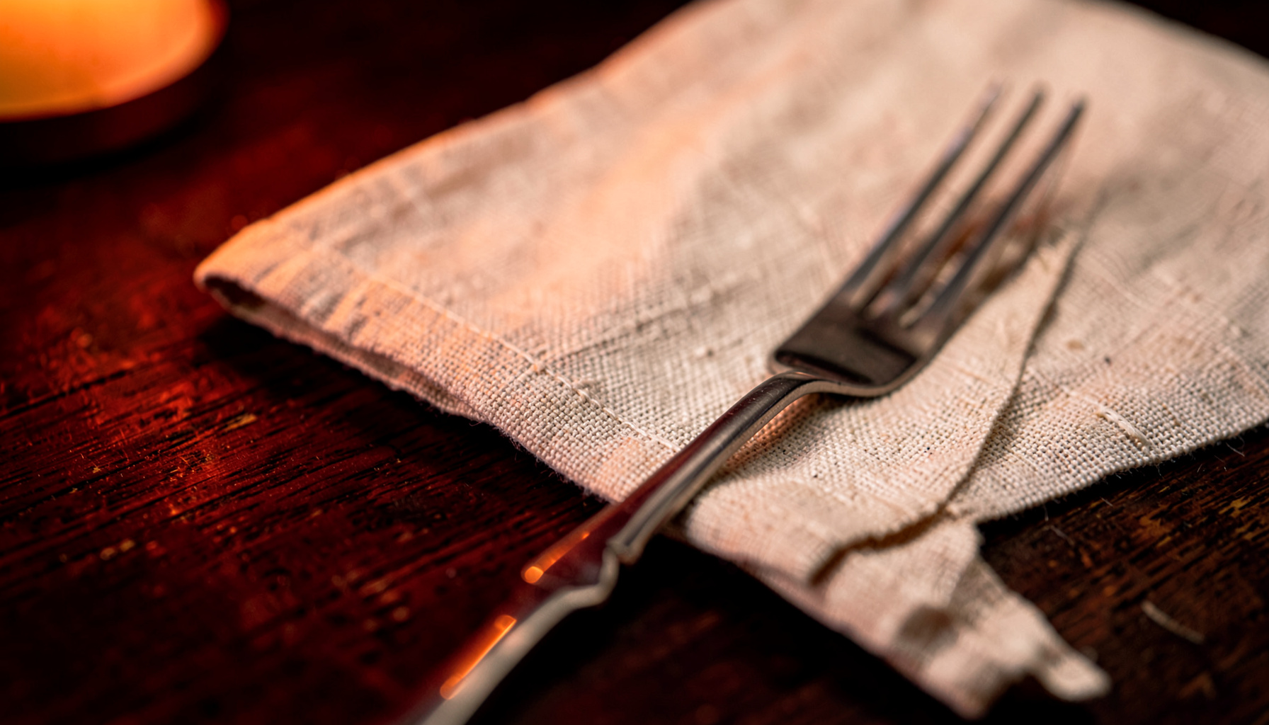 Extreme close-up photograph of a textured linen napkin beside a polished fork on a dark wooden tabletop, warm candlelight glow, deep browns and soft cream tones