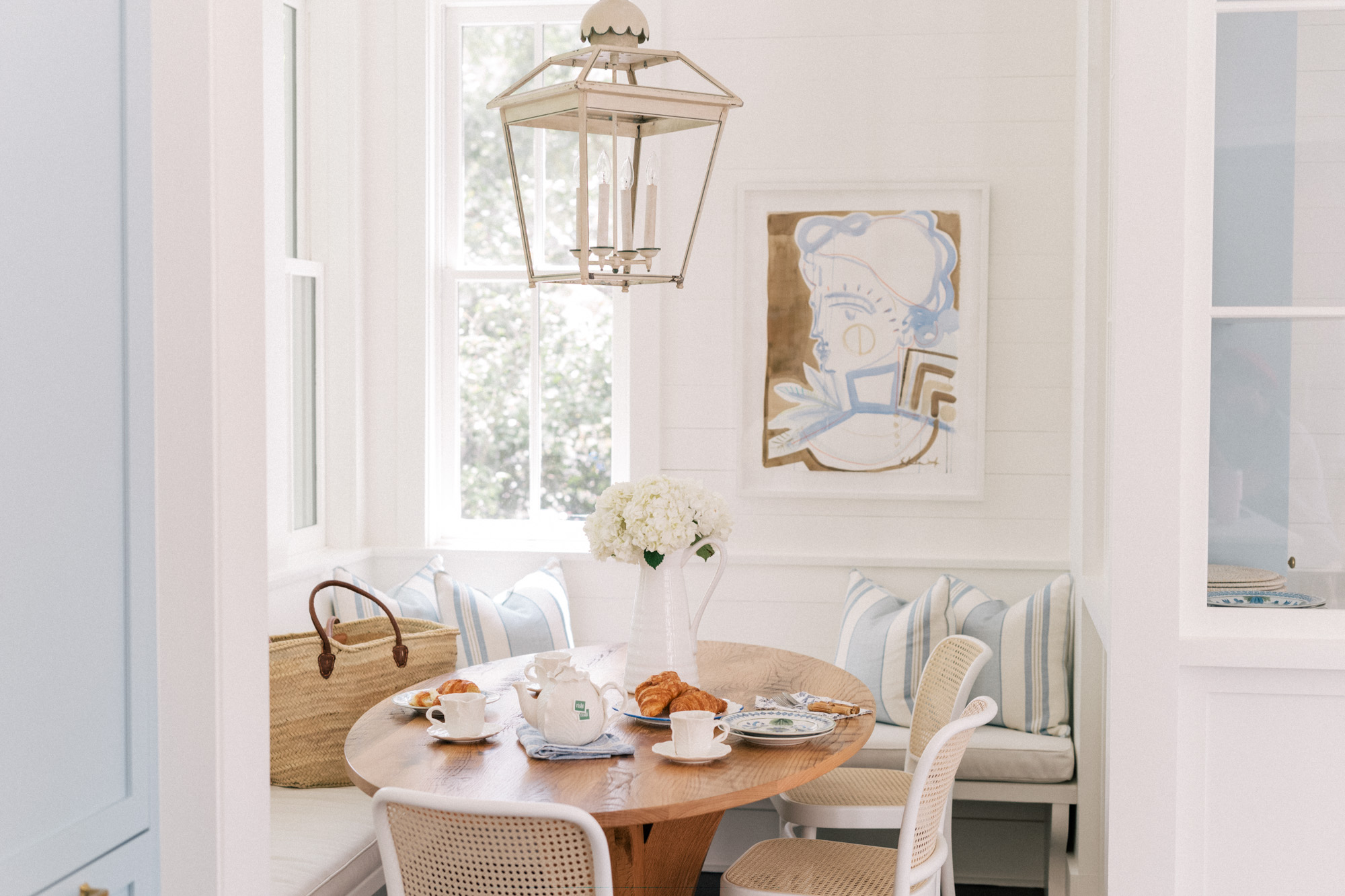Bright breakfast nook with round wooden table, striped pillows, pitcher of flowers, and pastries, light and airy mood